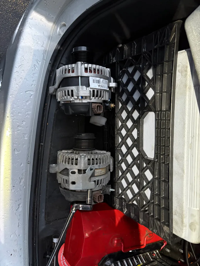 Two alternators side by side on a vehicle grille in the rain, one removed and one new