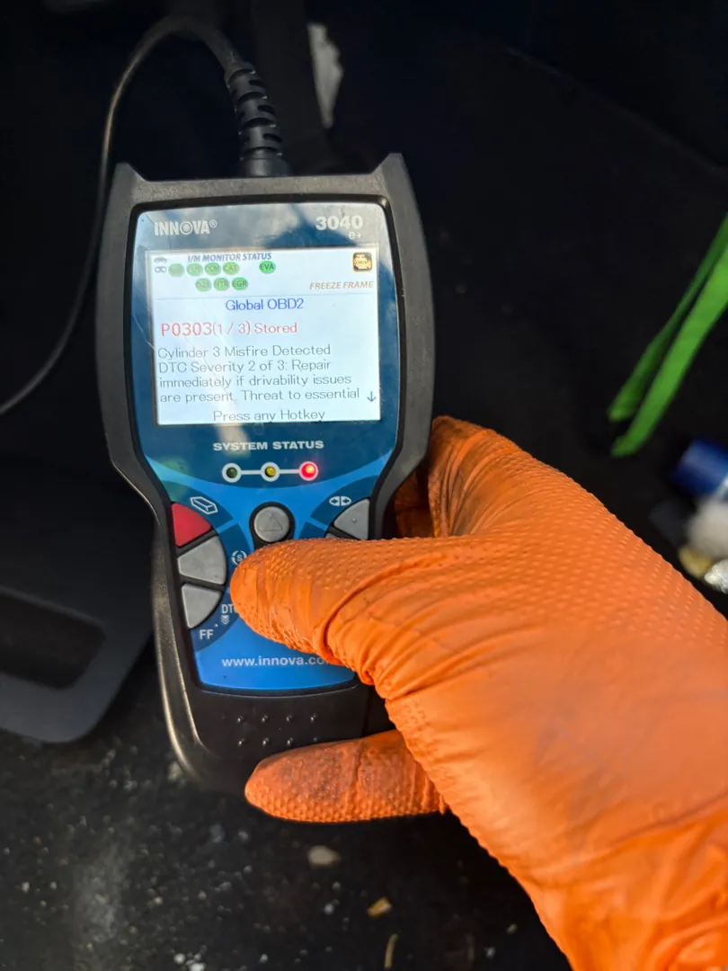 Handheld OBD2 scan tool held in an orange nitrile glove showing fault code P0303, cylinder 3 misfire detected